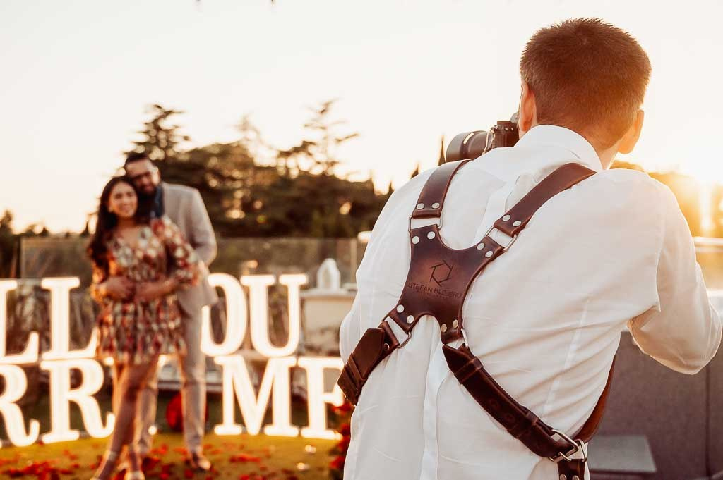 Fotografo di matrimonio al lavoro durante il tramonto, con una coppia felice sfocata sullo sfondo, accanto a decorazioni con la scritta 'Just Married' che creano un'atmosfera romantica e festiva.