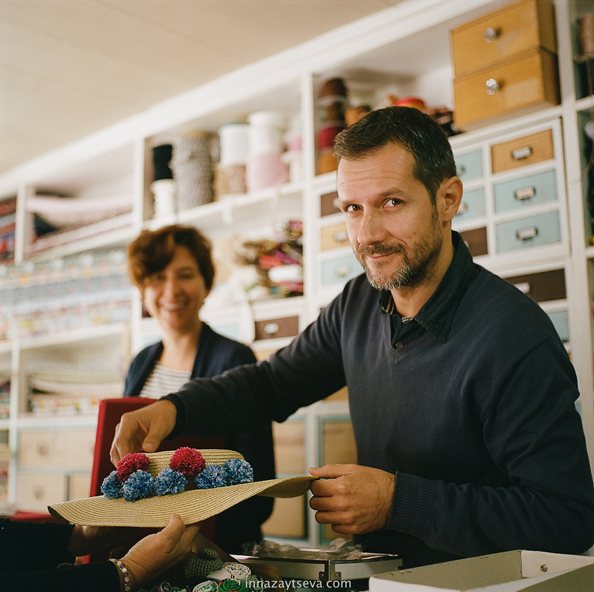man is fixing the hat in his workshop studio full of lockers with material and his female colleague on the background