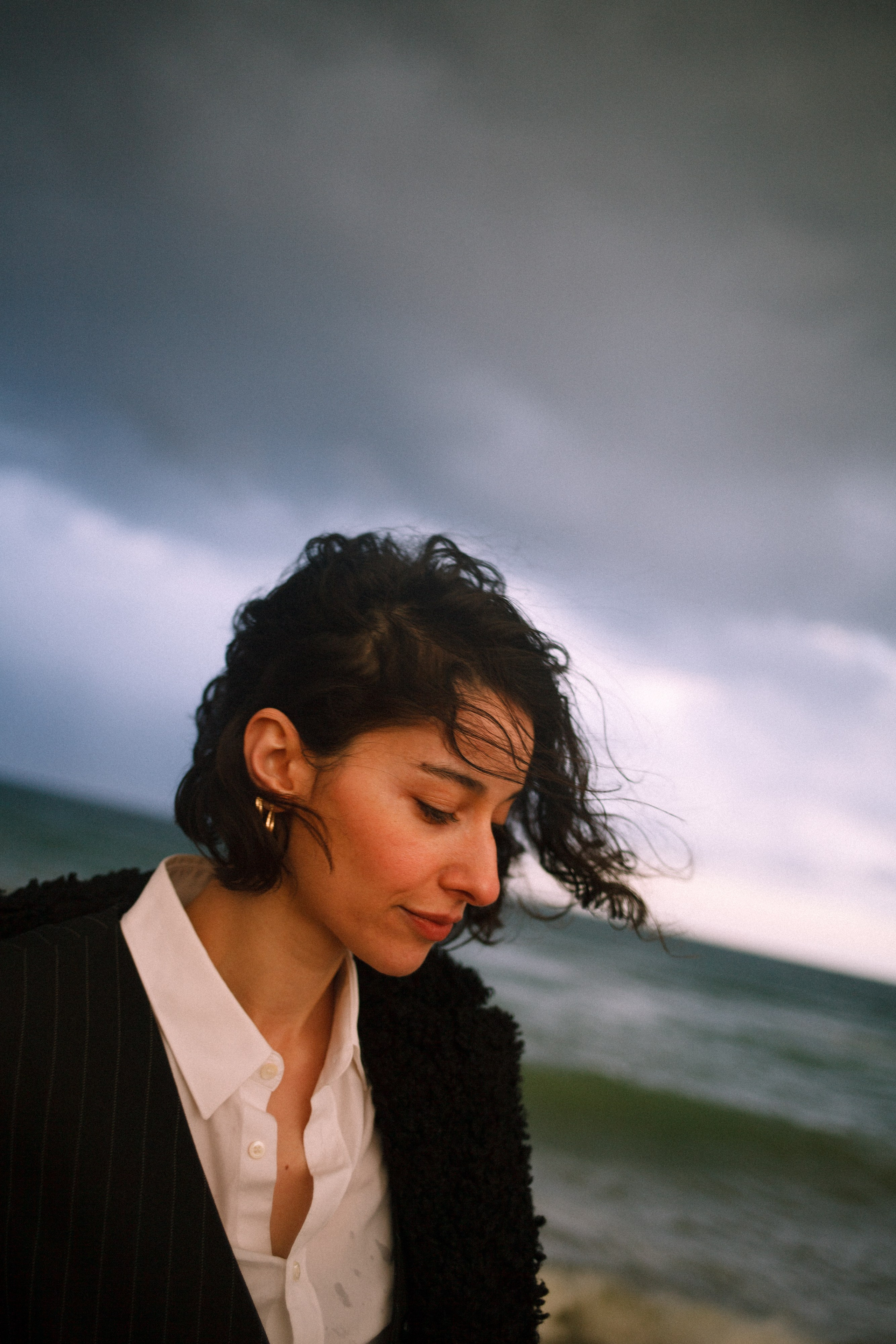 Woman with curly hair wearing a white shirt and black jacket, looking down with a cloudy sky and sea in the background