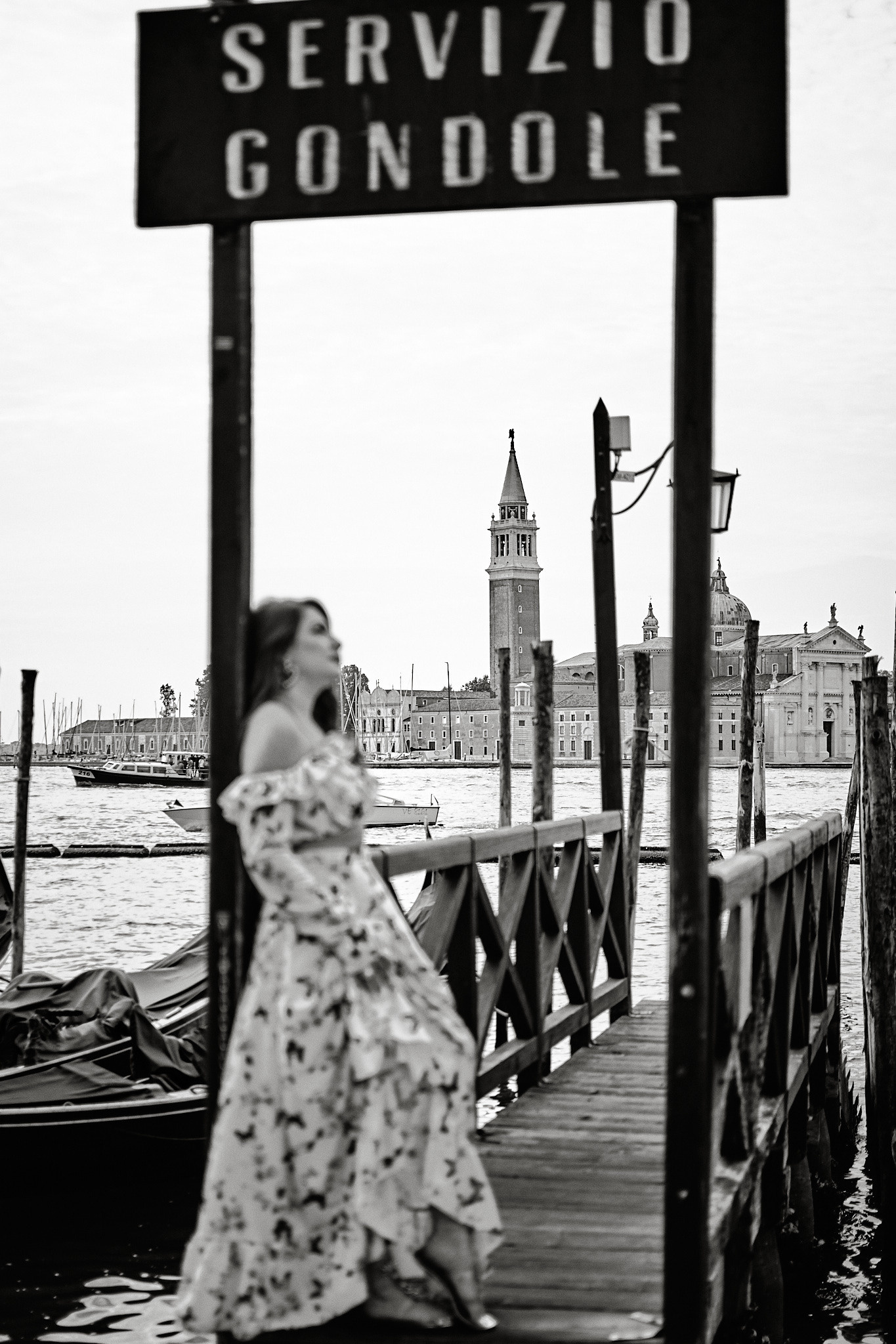 Girl in flowing dress standing on pier at St. Mark's Square with a clear view of the lagoon in the background. The girl's silhouette is out of focus. Black and White Photo - Venice photographer - travel blogger photography - Fashion Portrait photo - pier canal gondola lagoon B&W - Portrait shots in Venice, Italy 