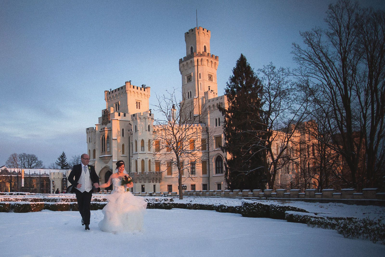 As the late evening sun casts its final glow, an English bride and groom are seen running across the snowy landscape of the historic Castle Hluboka.